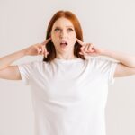 Portrait of stressed young woman covering her ears and gesticulating say no bla-bla-bla standing on white isolated background in studio.