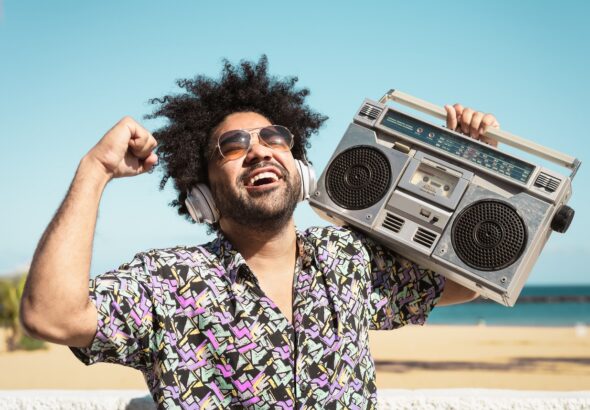 Happy Afro man having fun listening to music with wireless headphones and vintage boombox on the beach during summer time