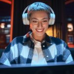 Young Beautiful Woman Wearing Headphones and Working from Home on Laptop Computer in Cozy Stylish Loft Apartment. Creative Female Smiling, Checking Social Media, Browsing Internet.