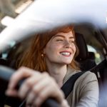 Woman Driving Car Smiling. Redhead Female Driver Behind the Wheel. Automotive Insurance, Safe Driving, and Commuting Concept. Young Woman in Modern Vehicle.