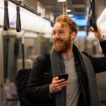 Happy male subway passenger is standing in the train wearing wireless headphones and a smartphone in his hand