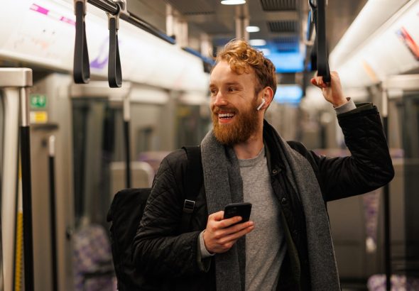 Happy male subway passenger is standing in the train wearing wireless headphones and a smartphone in his hand